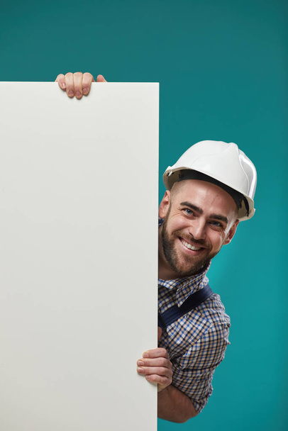 Vertical medium shot of happy Caucasian man in workwear and hardhat holding white blank poster looking at camera smiling - Foto, imagen