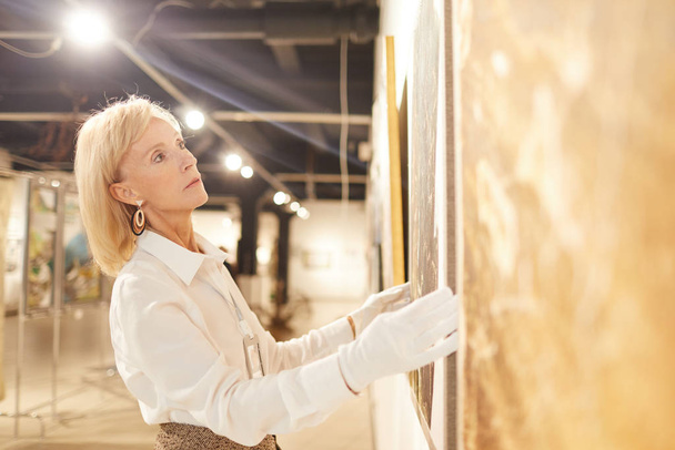 Side view portrait of elegant mature woman hanging paintings while working in art gallery or museum, copy space - Fotó, kép
