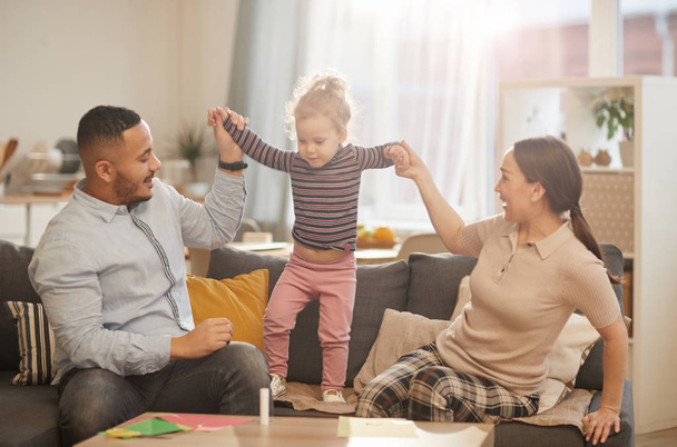 Caliente retrato tonificado de la familia moderna feliz jugando con la pequeña hija linda en el acogedor interior del hogar
 - Foto, Imagen
