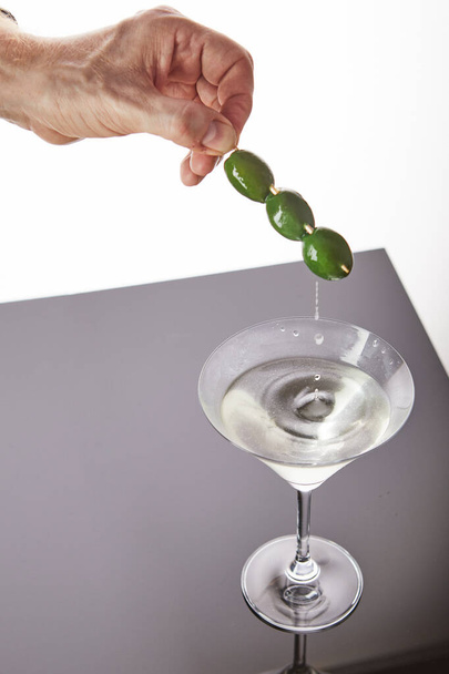 A glass with martini and a hand holding olives on a stick, drops flowing from olives into a glass on a gray white background - Photo, Image