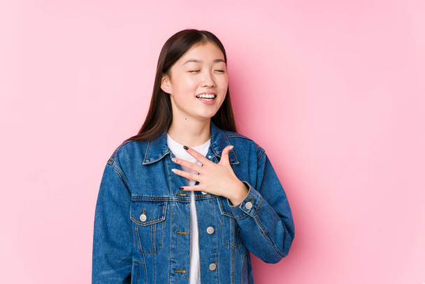 Jovem chinesa posando em um fundo rosa isolado ri em voz alta mantendo a mão no peito
. - Foto, Imagem