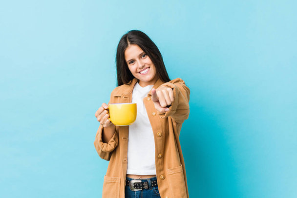 Молодая белая женщина, держа в руках кофейную кружку, весело улыбается, указывая на фронт.
. - Фото, изображение