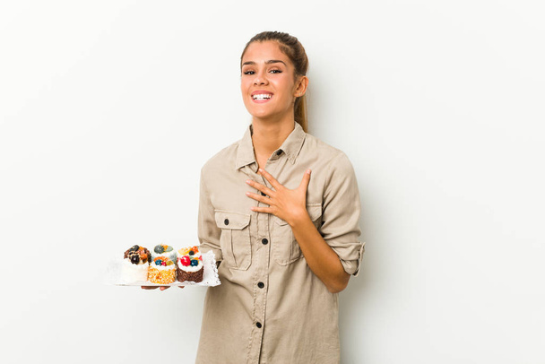 Joven mujer caucásica sosteniendo dulces pasteles se ríe en voz alta manteniendo la mano en el pecho
. - Foto, Imagen