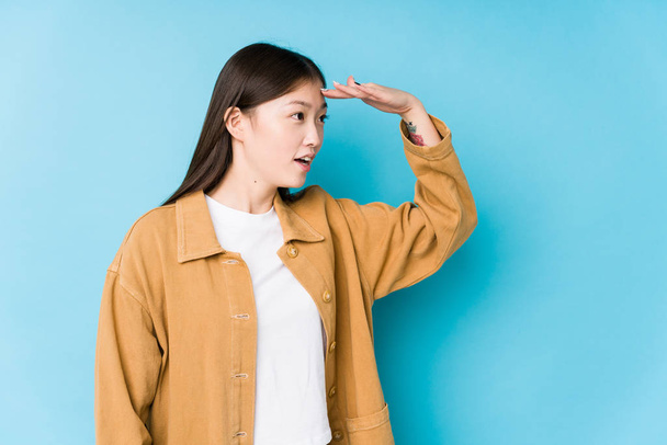 Mulher chinesa jovem posando em um fundo azul isolado olhando para longe mantendo a mão na testa
. - Foto, Imagem