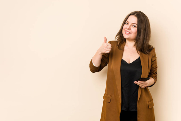 Jonge bochtige vrouw met een telefoon glimlachend en opheffen duim omhoog - Foto, afbeelding