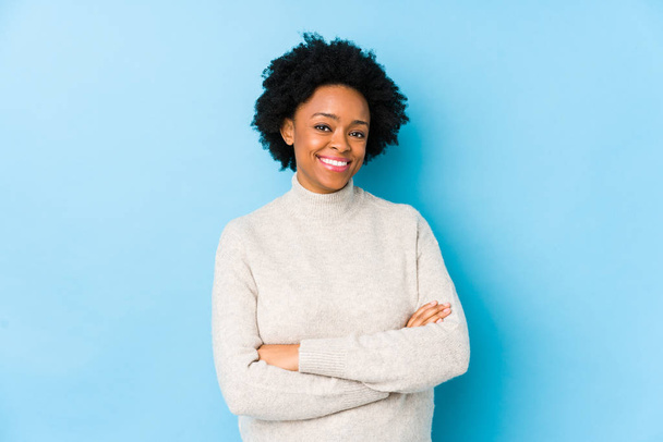 Afro-Amerikaanse vrouw van middelbare leeftijd tegen een blauwe achtergrond geïsoleerd die zich zelfverzekerd voelt, armen overstekend met vastberadenheid. - Foto, afbeelding