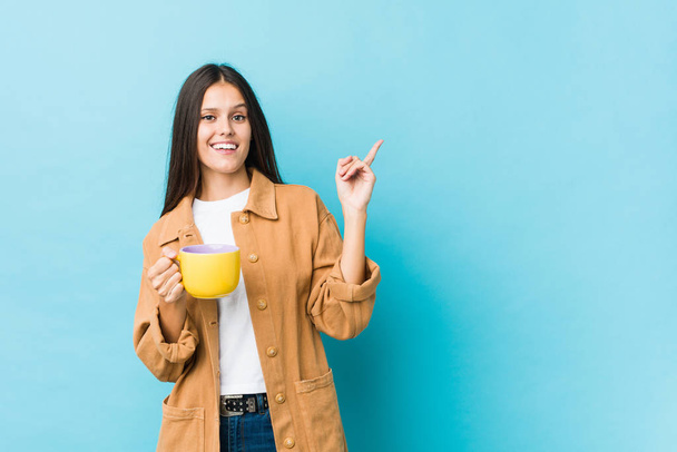 Joven mujer caucásica sosteniendo una taza de café sonriendo alegremente señalando con el dedo índice de distancia
. - Foto, Imagen