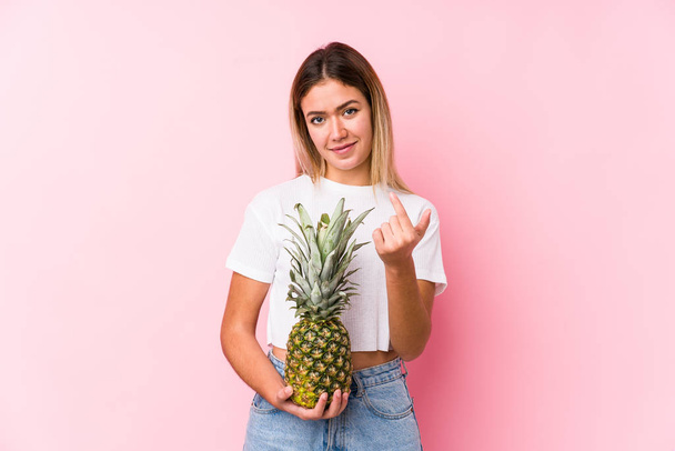 Młoda kaukaska kobieta trzyma ananasa wskazując palcem na ciebie, jakby zapraszając zbliżyć. - Zdjęcie, obraz