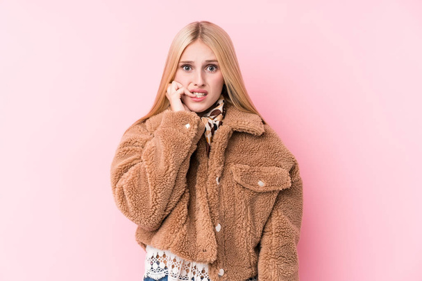 Jonge blonde vrouw draagt een jas tegen een roze achtergrond bijtende nagels, nerveus en zeer angstig. - Foto, afbeelding