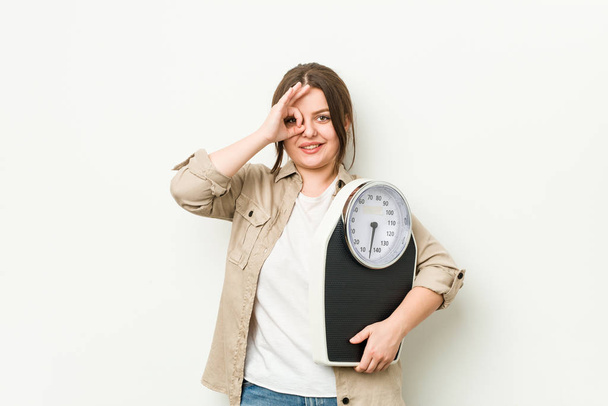 Young curvy woman holding a scale excited keeping ok gesture on eye. - Photo, image