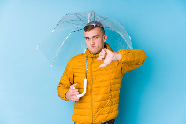 Młody biały mężczyzna z parasolem, okazujący niechęć do gestów, opuszczone kciuki. Koncepcja sporu. - Zdjęcie, obraz