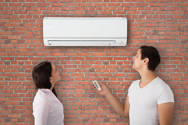 Young Couple Switching On The Air Conditioner With Remote Control Attach On Brick Wall - Foto, immagini