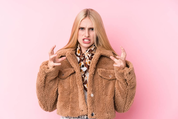 Young blonde woman wearing a coat against a pink background upset screaming with tense hands. - 写真・画像