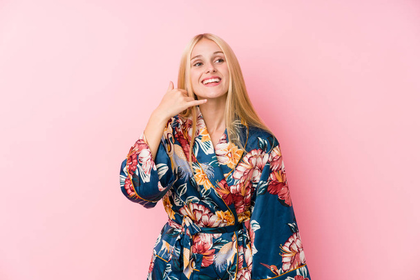 Jeune femme blonde portant un pyjama kimono montrant un geste d'appel téléphonique mobile avec les doigts
. - Photo, image