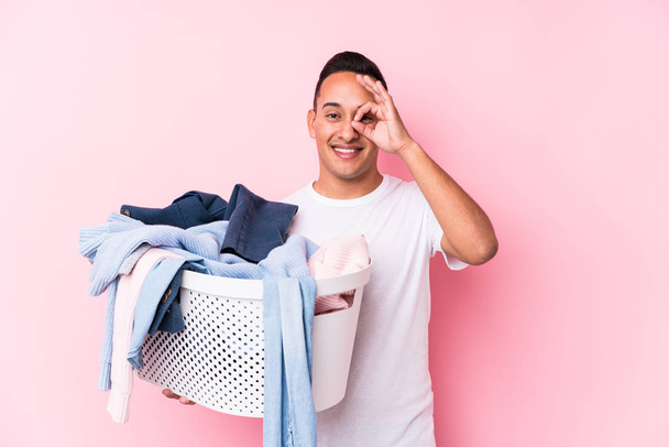 Giovane uomo latino raccogliendo vestiti sporchi isolato eccitato mantenendo ok gesto sugli occhi
. - Foto, immagini
