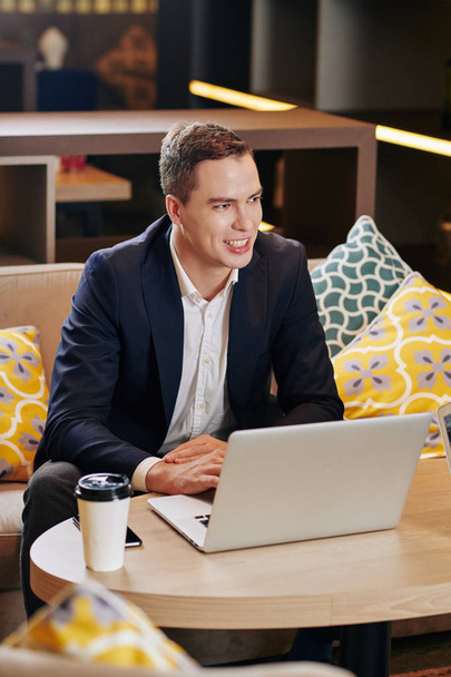 前向きなハンサムな白人ビジネスマンがコーヒーを飲みながらカフェテーブルでノートパソコンで作業する - 写真・画像