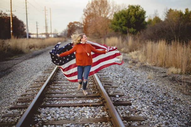 Happy white female holding the flag of the United States of America running on the railway - Foto, Bild
