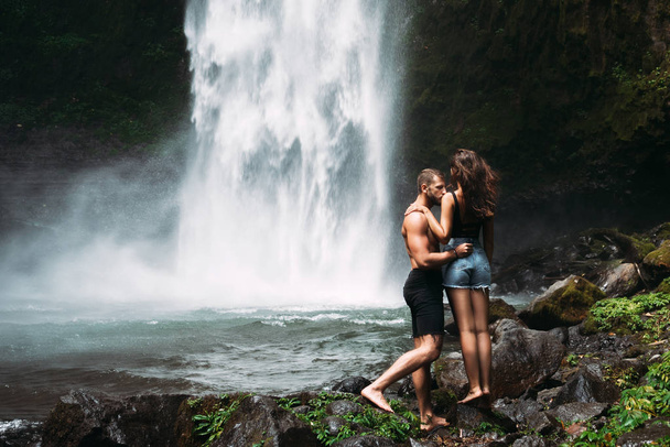 A man of athletic build kisses a beautiful girl at the waterfall. Honeymoon trip. Declaration of love. A couple in love on a waterfall. Happy couple in Bali. Beautiful couple travels the world - Foto, Bild