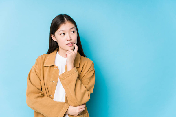 Young chinese woman posing in a blue background isolated relaxed thinking about something looking at a copy space. - Photo, Image