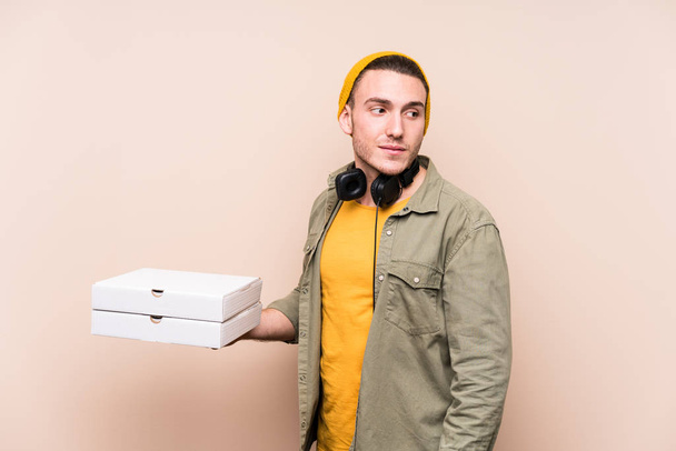 Молодой кавказский мужчина с пиццей смотрит в сторону улыбаясь, веселый и приятный
. - Фото, изображение
