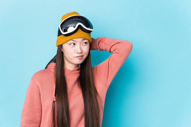 若い中国の女性は、頭の後ろに触れる隔離されたスキー服を着て、考え、選択をする. - 写真・画像