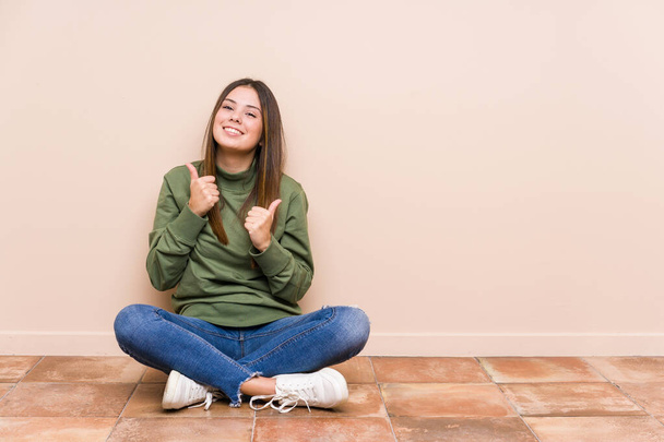Fiatal fehér nő ül a padlón elszigetelt emeli mindkét hüvelykujját fel, mosolygós és magabiztos. - Fotó, kép