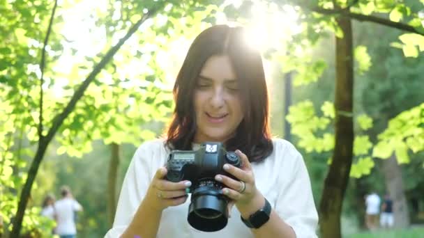 Pretty young girl laughs loudly while browsing photos on professional photo camera. Pretty smiling and laughing girl photographer wearing white shirt takes photos in a park. - Footage, Video