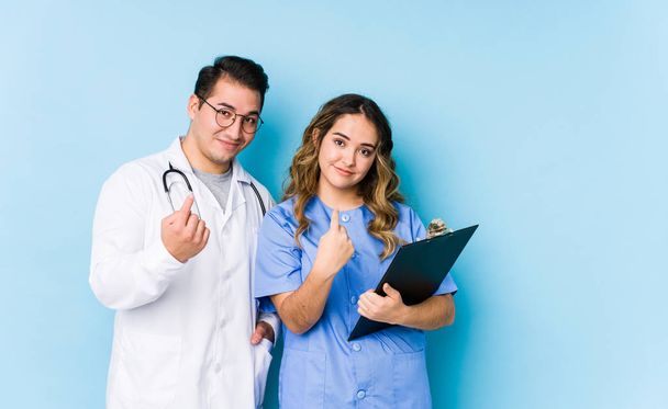 Joven pareja de médicos posando en un fondo azul aislado señalándote con el dedo como si invitaras a acercarte
. - Foto, Imagen