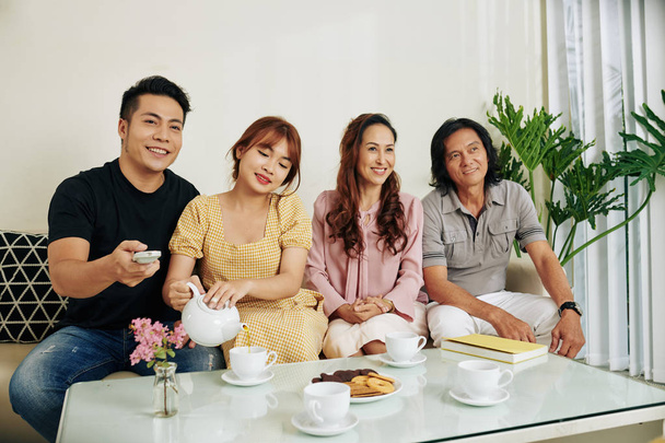 Feliz familia asiática con niños adultos tomando té con galletas y viendo la televisión en casa
 - Foto, Imagen