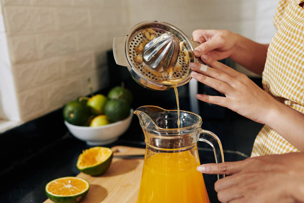 Руки женщины наливают свежий апельсиновый сок из металлического ручного сжимателя в стеклянную банку
 - Фото, изображение
