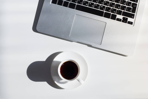 Cup of coffee and laptop computer on white table, top view. Simple minimalistic flat lay of modern workplace or simplistic clean office desk, pc with cyrillic keyboard - Photo, Image