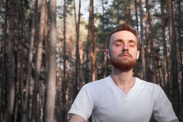 森の中で近代的な技術を使って瞑想している若者。松の木に座って屋外で瞑想を楽しむアクティブな男. - 写真・画像