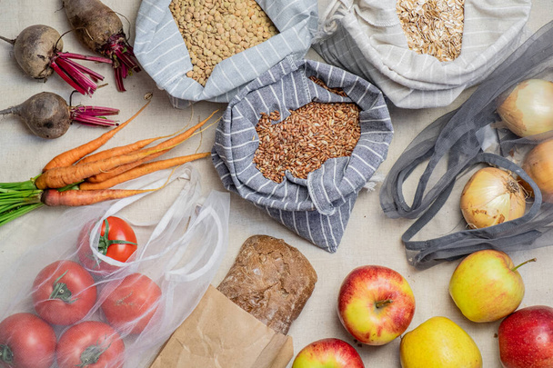 Groceries in reusable bags on natural flax or hemp background, top view. Concept of zero waste ethical shopping: raw vegan food including fruit, vegetables, rice and cereal in bio packaging, flat lay view - Photo, Image