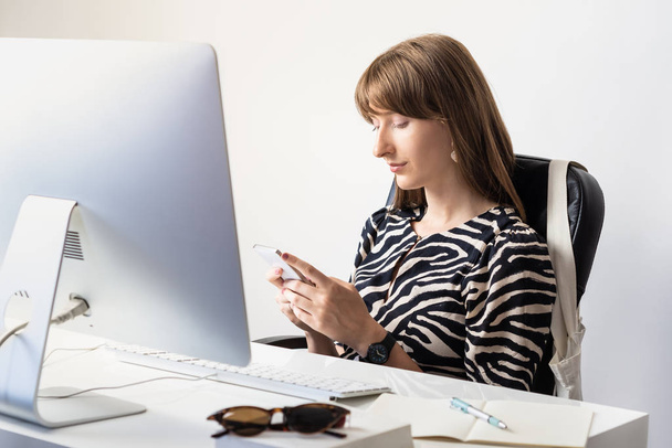 Young female worker at desk in modern minimalistic office. Portrait of business woman using mobile phone in front of desktop computer - 写真・画像