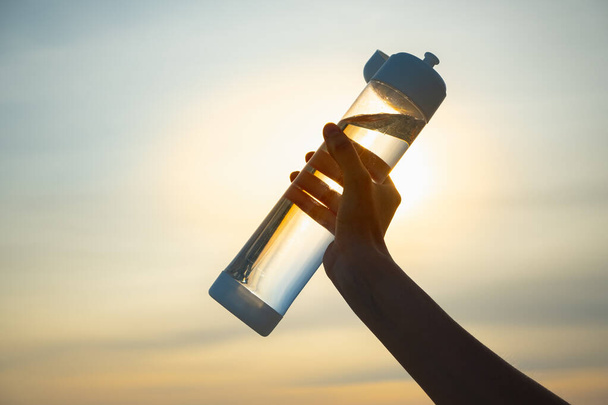 Human hand holds a water bottle against the setting sun. Close up of a reusable water bottle in a human hand, concept of thirst, rehydration and decreasing single use plastic - Fotografie, Obrázek