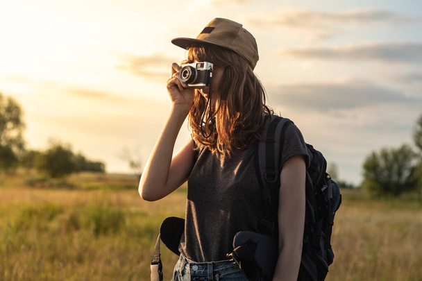 Young female tourist taking photo at hiking trip. Woman with backpack stands at sunset and photographs beautiful rural area - Φωτογραφία, εικόνα