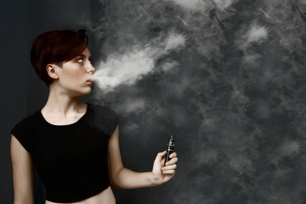young girl smokes electronic cigarette. female model vaping a vaporizer on black metal wall background. face of vaping young woman at black studio. no smoking day          - Foto, Imagen