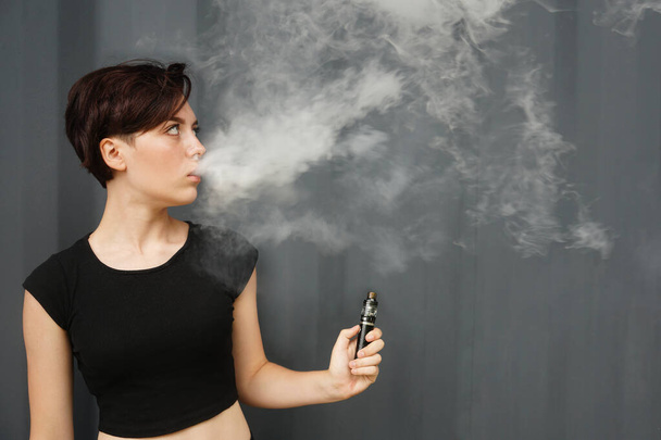 young girl smokes electronic cigarette. female model vaping a vaporizer on black metal wall background. face of vaping young woman at black studio. no smoking day          - Foto, Bild