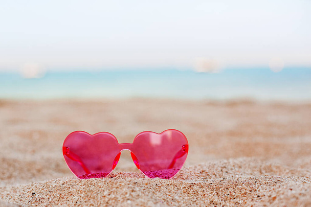 Vaaleanpunaiset aurinkolasit hiekkarannalla, jossa on sininen meritausta, häämatka, loma, kopiotila
 - Valokuva, kuva