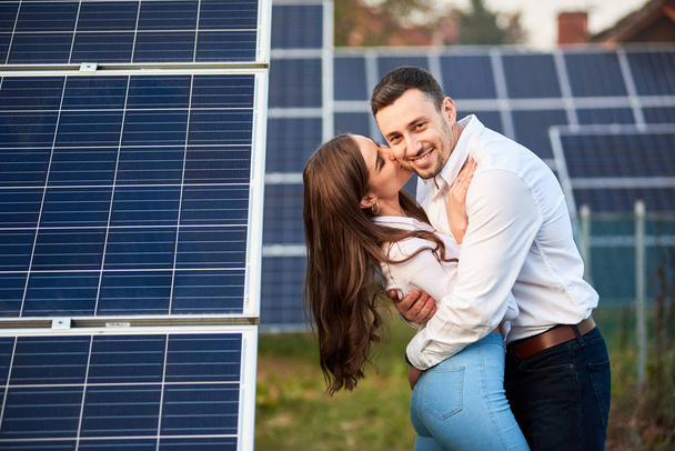Happy pair halaavat taustaa vasten rivi aurinkopaneelit sivuston lähellä taloa. Pitkätukkainen tyttö suutelee miestä. Hoikkia farkuissa ja valkoisissa paidoissa. Aurinkoenergia käsite kuva
 - Valokuva, kuva