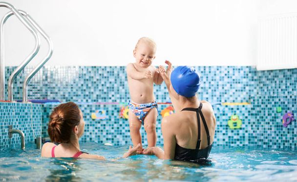 Back view of two women in swimming pool helping a small boy keep balance in water, happy baby is standing in their hands. Concept of sport for kids - Photo, Image