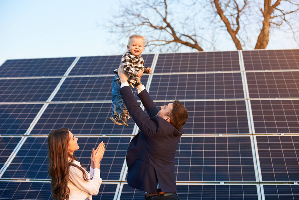 Tiefansicht einer glücklichen Familie, die ihre Zeit in einer Solaranlage verbringt, Vater hält seinen Sohn über ihren Köpfen, Mutter klatscht in die Hände - Foto, Bild