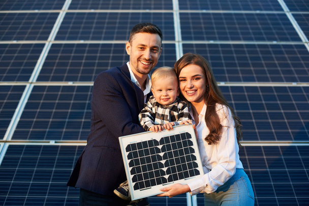 Portrait of a young family holding a small solar panel and a baby boy, smiling and looking at camera. Concept of green generation - Photo, Image