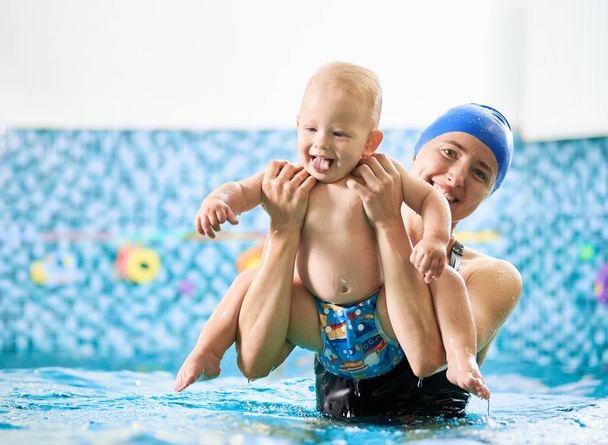 Young smiling woman is keeping her baby boy over turquoise water in the pool doing physical exercises, playful child is showing his tongue. Concept of early development - Photo, Image