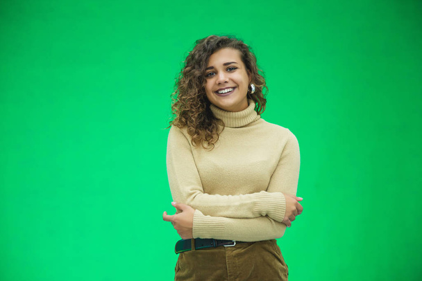 Portrait of a beautiful friendly african american woman with curly hairstyle and lovely smile isolated on a green background, her hands folded. - Photo, Image