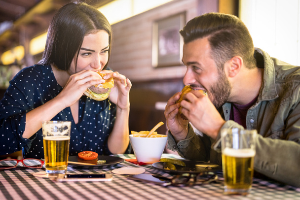 Happy couple having fun eating burger at restaurant pub fast food - Young people enjoying moment at indoor diner location - Relationship concept with focus on girl face with warm bivid filter - Fotografie, Obrázek