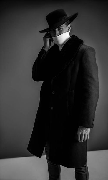 Mode-Modell posiert auf grauem Hintergrund im Studio. Modellversuche. Mann mit schwarzem Mantel, weißem Golf und Hut. - Foto, Bild