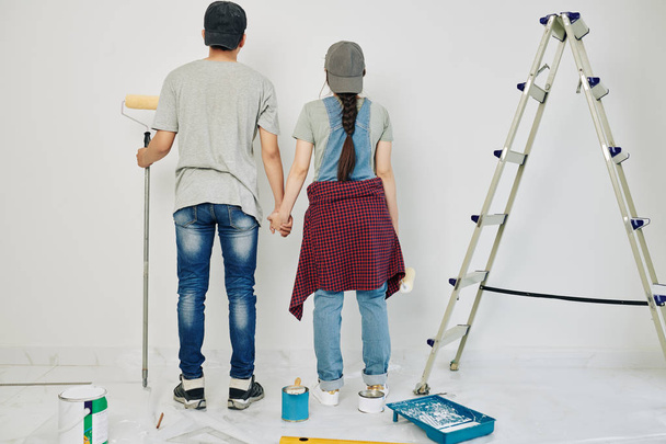 Jeune couple tenant la main et regardant le mur qu'ils vont peindre, vue de dos
 - Photo, image