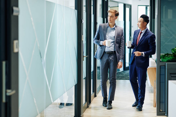 Gente de negocios multiétnicos caminando por el pasillo de la oficina con tazas de café y discutiendo noticias
 - Foto, imagen