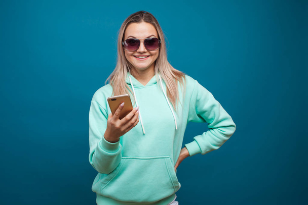 Cheerful blonde with a mobile phone. Portrait of a young positive woman in a blue hoodie - Foto, Imagem